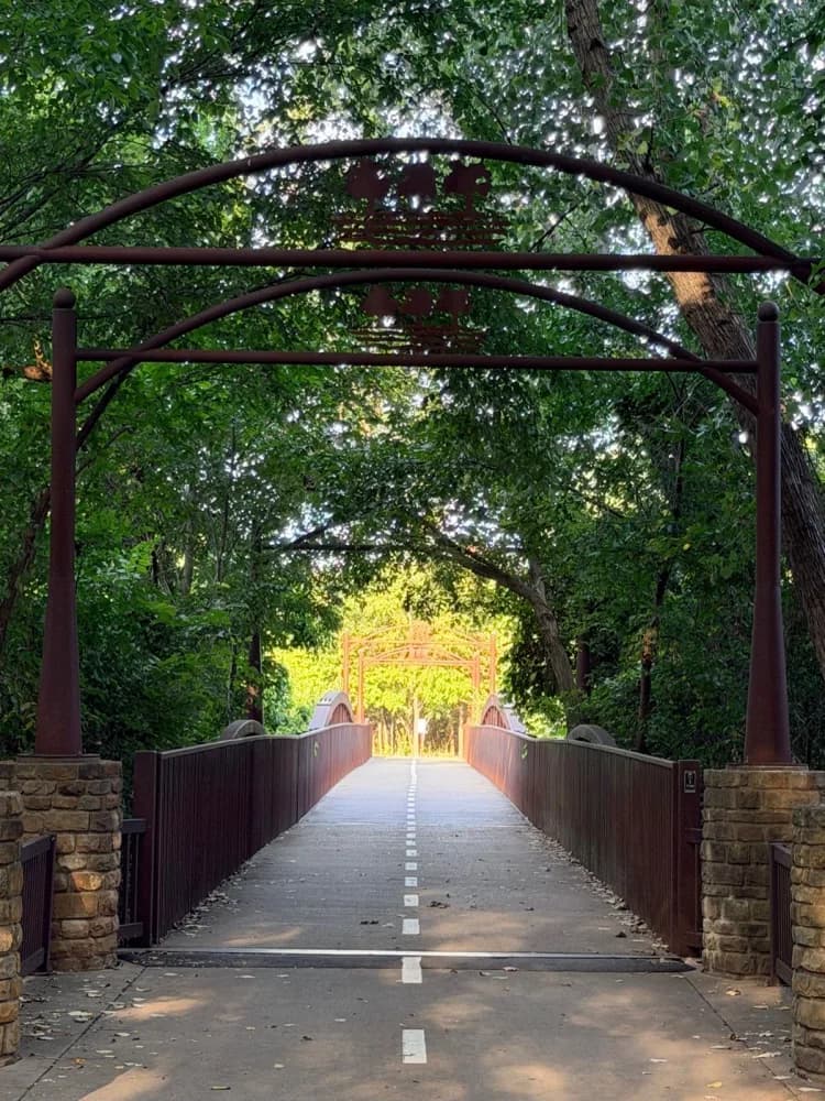 A steel arch footbridge into green trees on a Fort Worth trail