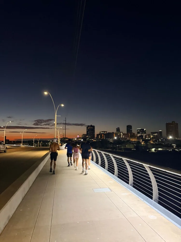 Runners on a bridge at dusk with the Fort Worth skyline lit behind them